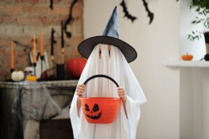 Halloween candy exposure in FBT for eating disorders [Image description: child in a ghost costume with a witches hat holding a plastic pumpkin pail filled with candy] Represents a teen in recovery from an eating disorder who is enjoying candy on Halloween with parental support