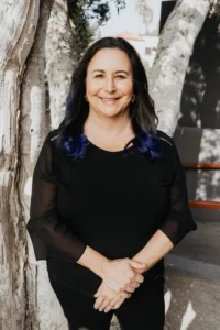 Lauren Muhlheim, Psy.D. [Image description: photo of a middle-aged white woman with medium length black hair and a black shirt in front of a tree]