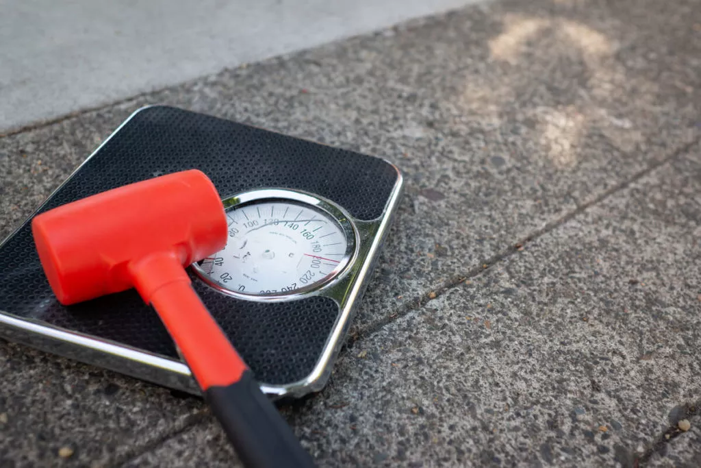 Accepting your body's setpoint [Image description: red hammer on a bathroom scale] represents the rejection of diet culture by a person in eating disorder recovery in Los Angeles, California