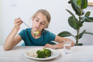 Feeding a Child with ARFID in Los Angeles, California [Image description: photo of a girl eating broccoli and rice and looking unhappy] Represents a child with ARFID in therapy in California