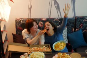 College Weight Gain in California [image description: 2 young women eating pizza joyfully with other snacks around] depicts potential college students in California who are not afraid of the Freshman 15