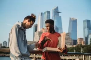 Supporting a friend with ARFID in California [Image description: 2 diverse men outside with a city scape behind them and one is holding a pizza box and offering pizza to the other man] Represents a person supporting a friend struggling with ARFID in California