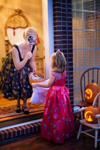 Halloween Candy and Intuitive Eating {Image description: a girl with a candy bag and wearing a costume with a pink dress is standing at the doorstep and an adult woman in costume is giving her candy] Represents a family in California who is practicing intuitive eating