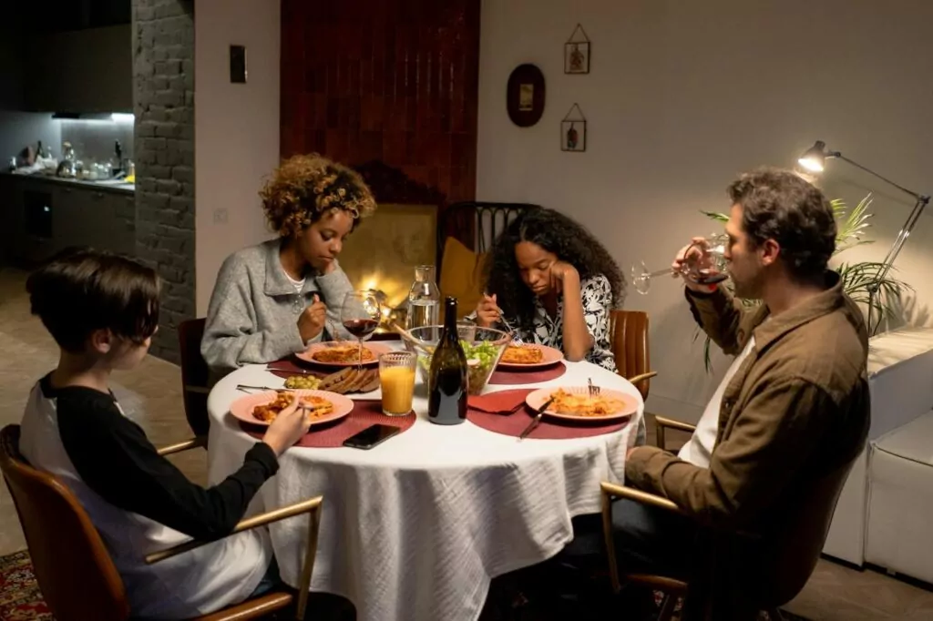 Family having dinner and unhappy in FBT [Image description: family of 2 adults and two kids and the teen girl looks very unhappy about eating] Represents a potential family in California doing FBT for their teen's eating disorder